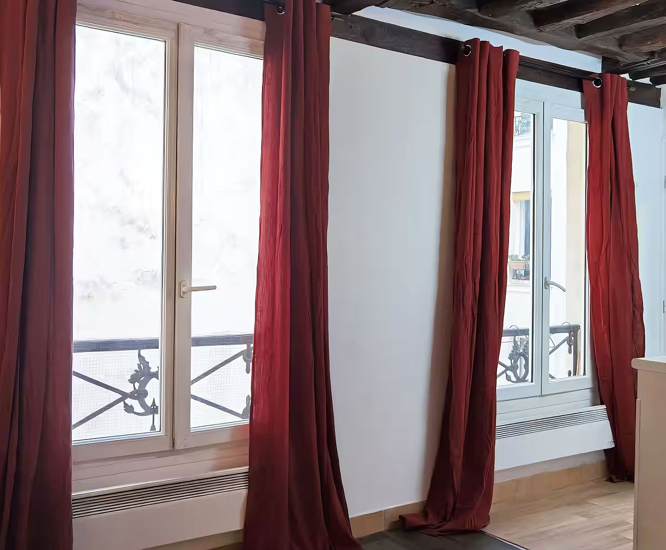 A room with large windows covered by red curtains, a wooden floor, and a visible balcony with a railing.