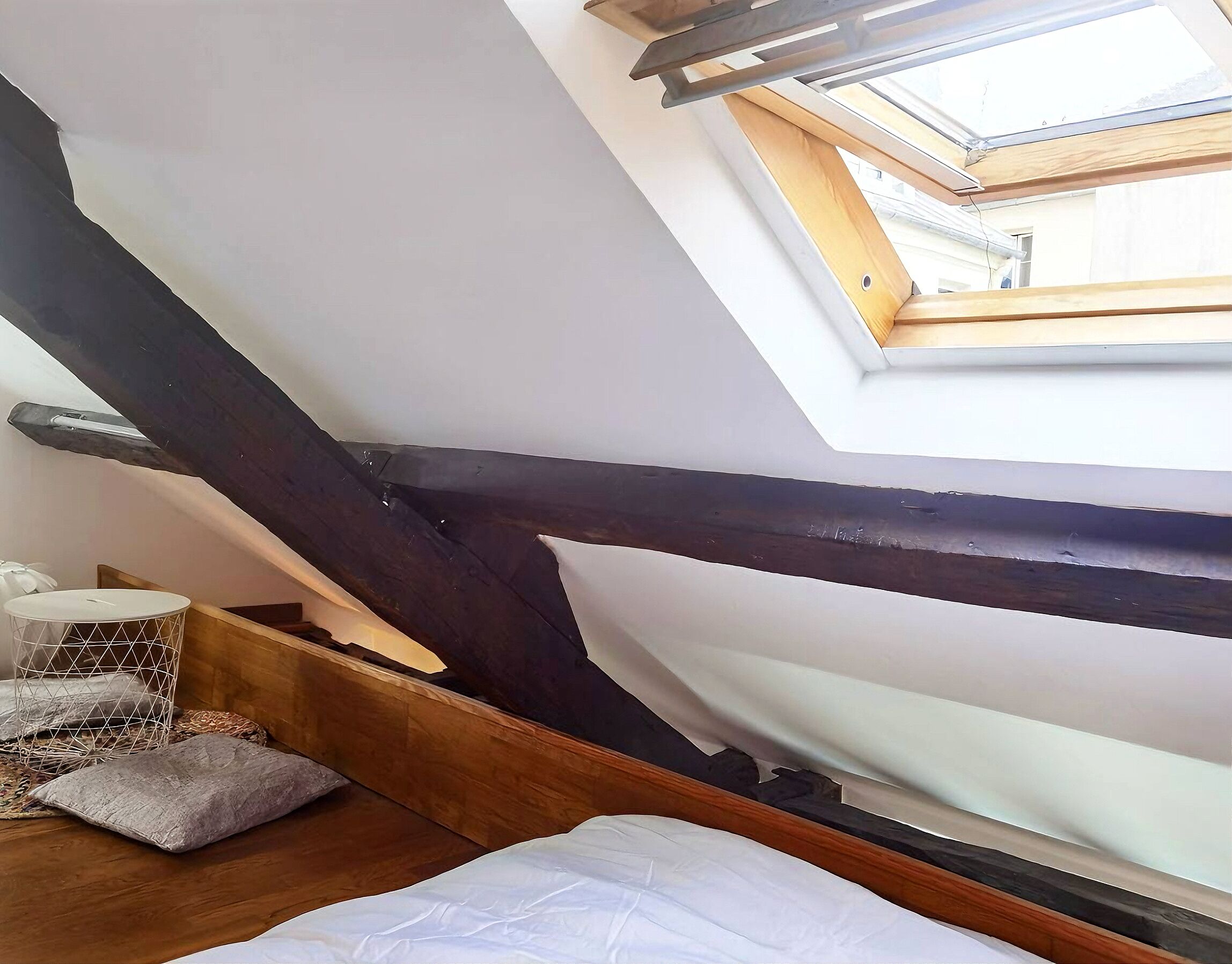 A sloped ceiling with a skylight, wooden beams, a white mattress, and a small white container.