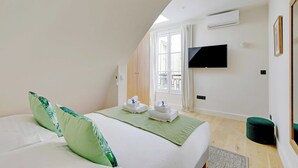 A hotel room with a bed, a green bedspread, a flat-screen TV mounted on the wall, and a window with curtains.