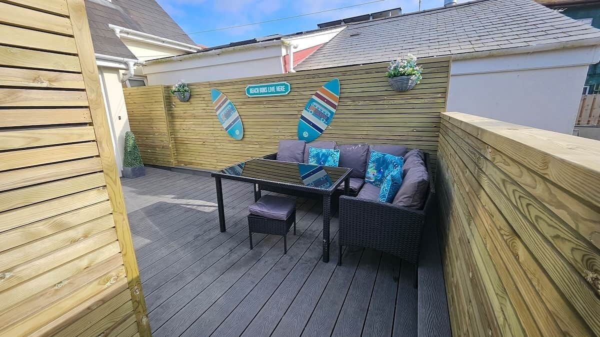 A wooden deck with a table and two chairs, a wooden fence, and a sign that reads 'BEACH HOURS LIVE HERE.'