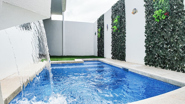 En swimmingpool med ett vattenfall, omgiven av vita väggar och grönskande växtlighet.