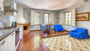Una cocina amplia con estufa a gas, armarios de madera y salpicadero de azulejos. Hay un comedor con mesa y sillas, y un sofá cubierto con un tejido azul.