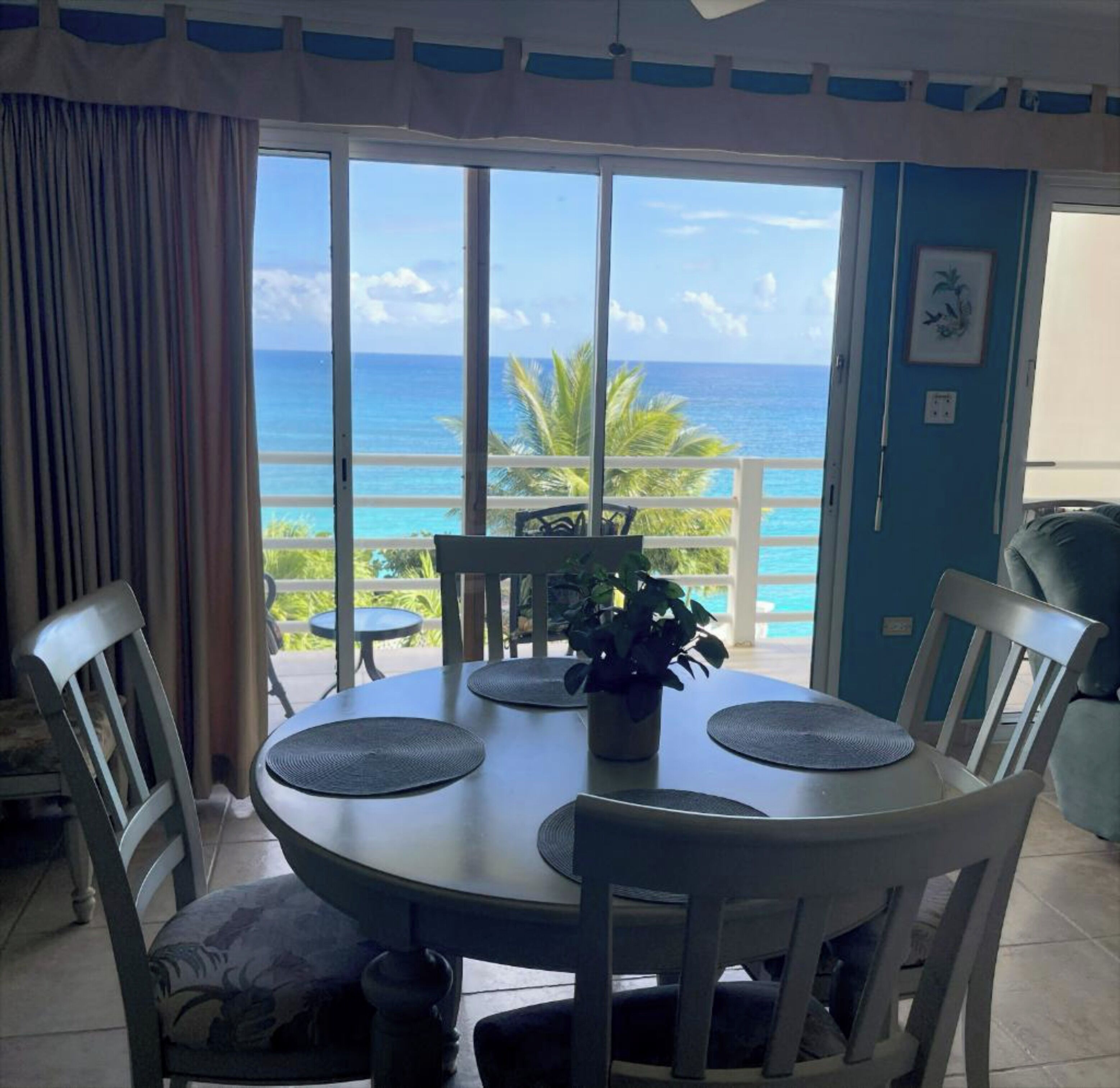 Un coin repas avec une table et des chaises, une plante en pot et une vue sur l’océan par de grandes portes vitrées.