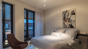 Een moderne slaapkamer met een groot bed, een gebogen fauteuil en een groot raam met zonwering.