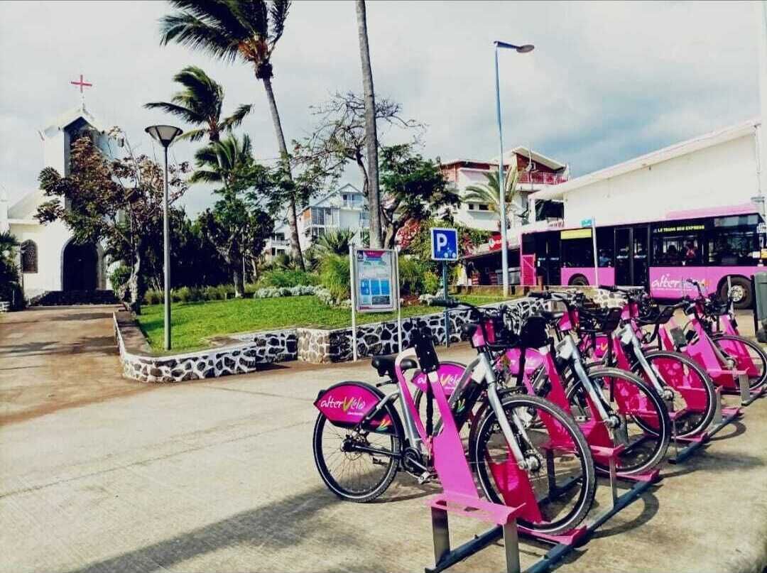 Une rangée de vélos roses est stationnée dans un espace prévu à cet effet, avec une église et un autobus en arrière-plan.