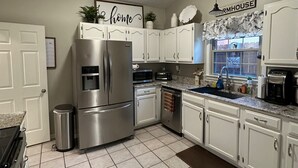 A kitchen with white cabinets, a stainless steel refrigerator, a sink, and a window with a 'Farmhouse' sign.