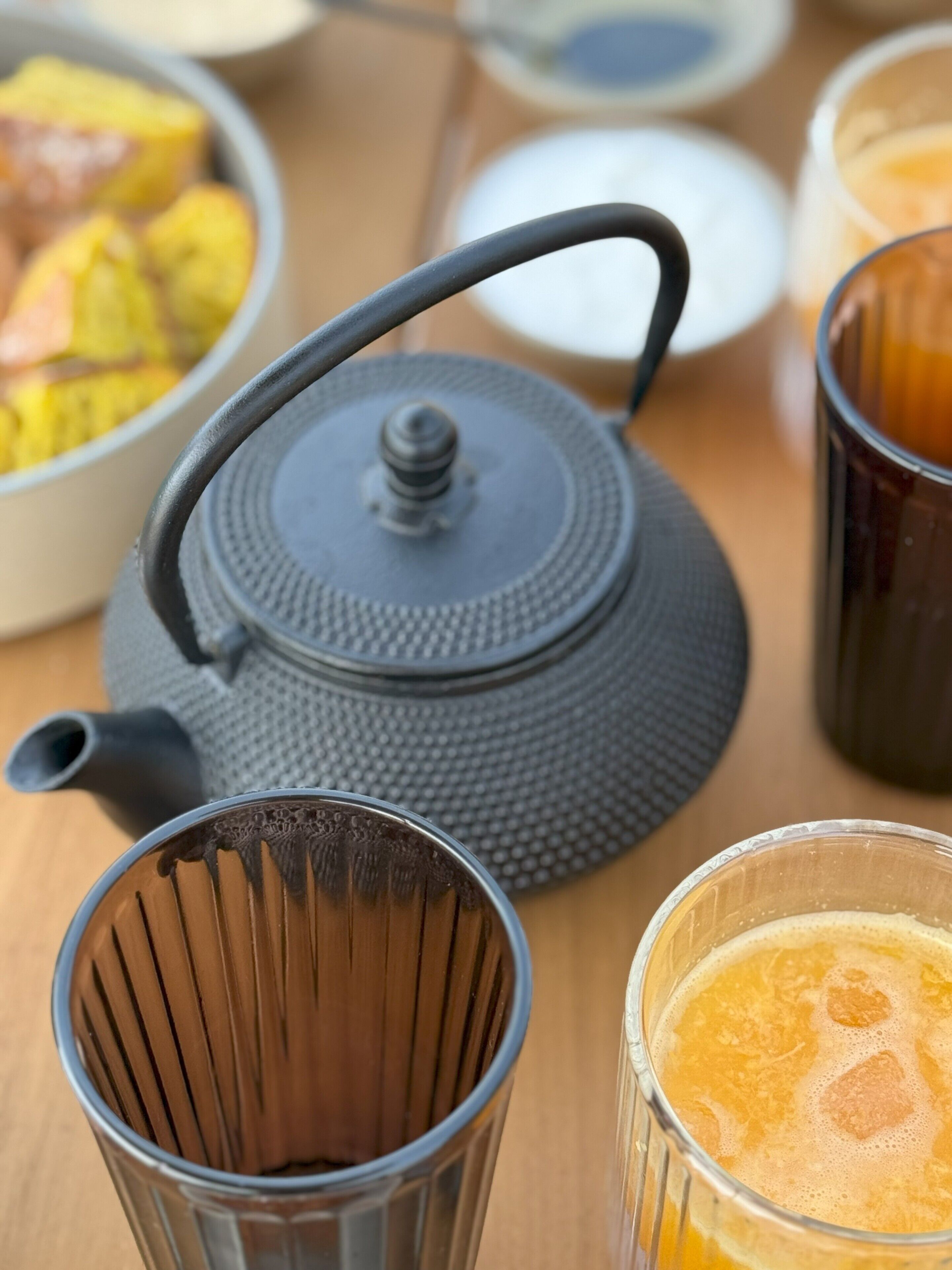 A teapot, a glass with a brown liquid, and a glass with a yellow liquid on a wooden table.