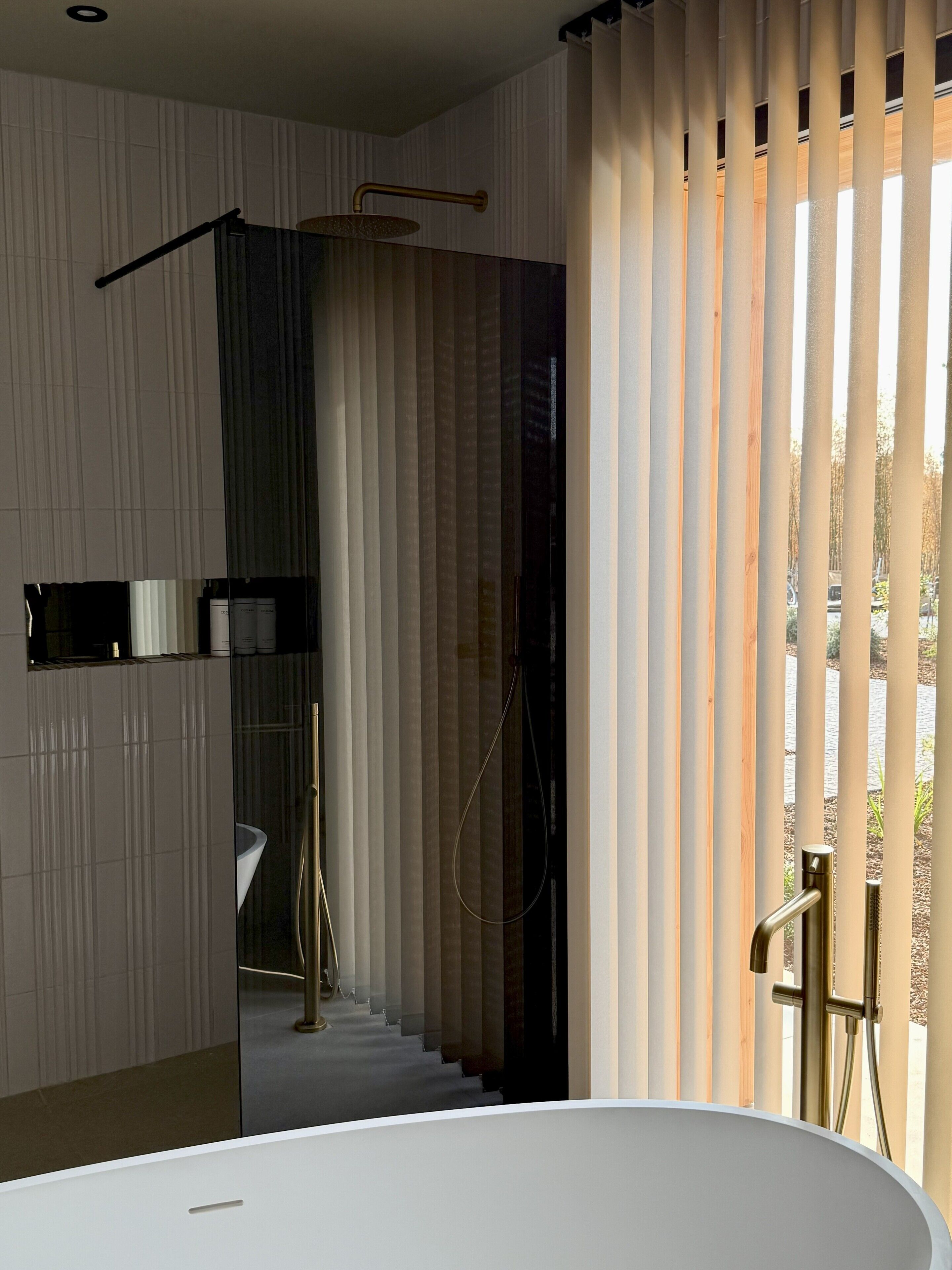 Ein modernes Badezimmer mit einer freistehenden Badewanne, einer Dusche mit goldener Armatur und Blick durch ein Fenster mit vertikalen Jalousien.
