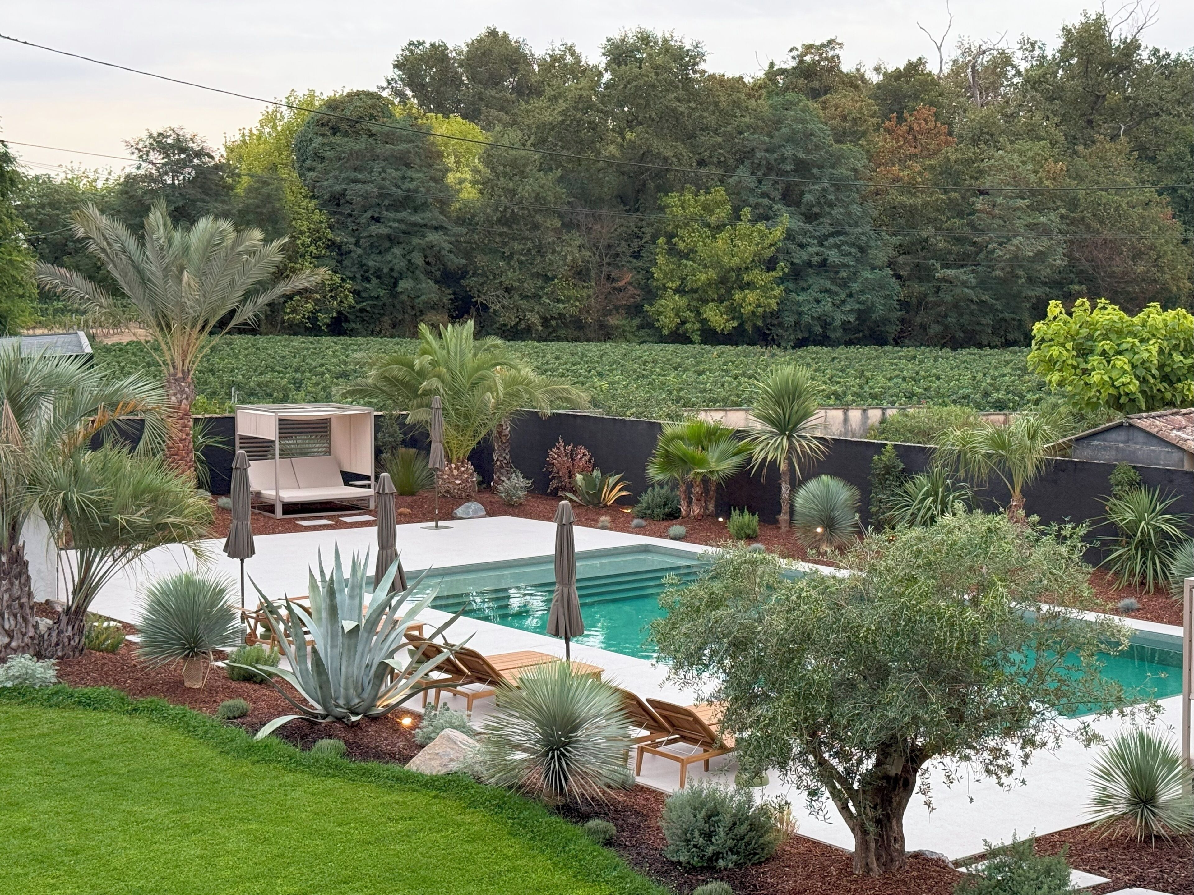 Un área de piscina con sillones de descanso y una pequeña terraza, rodeada de plantas y árboles.