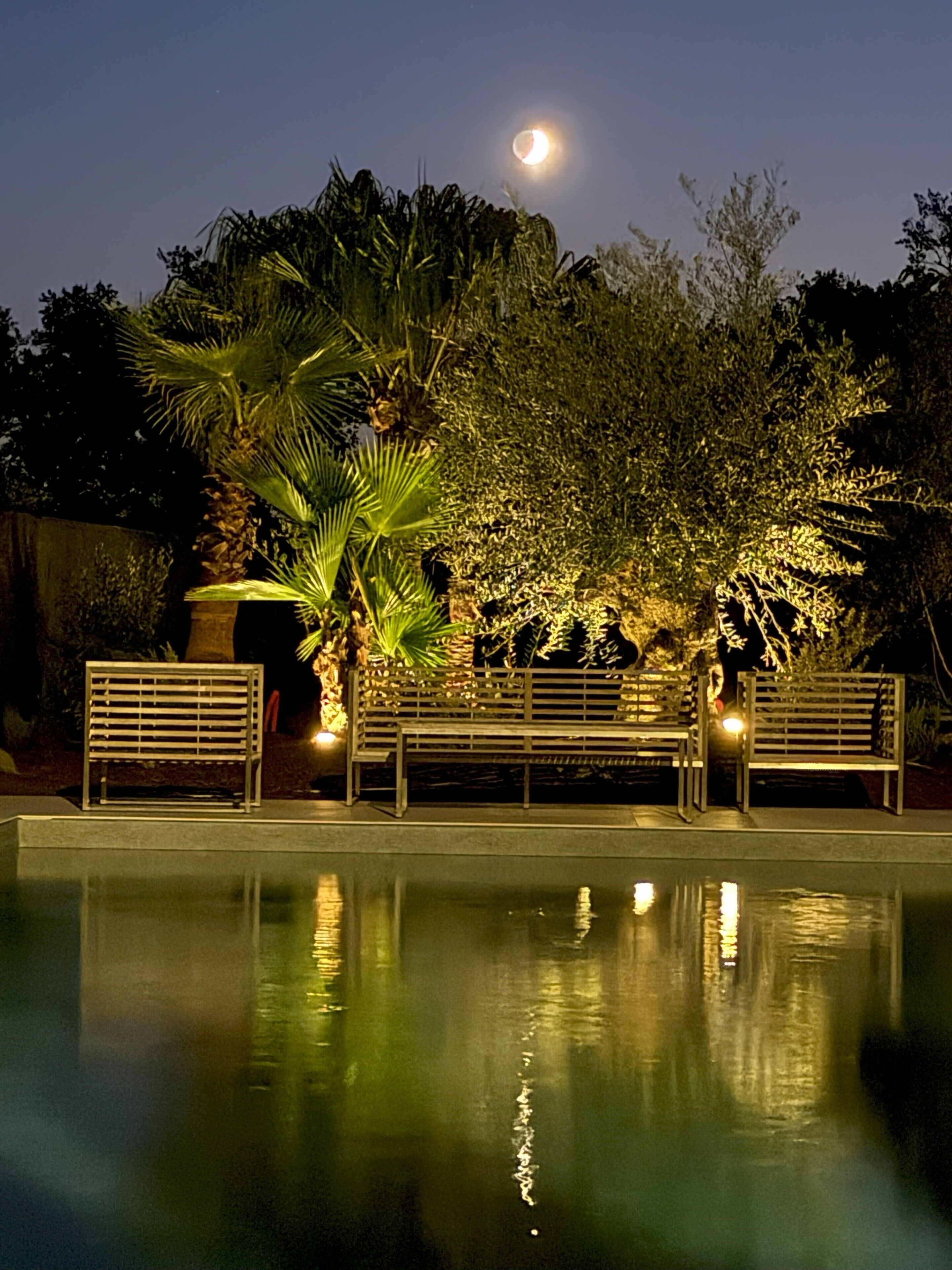 Een terras buiten bij nacht met banken en een zwembad dat de maan weerspiegelt.