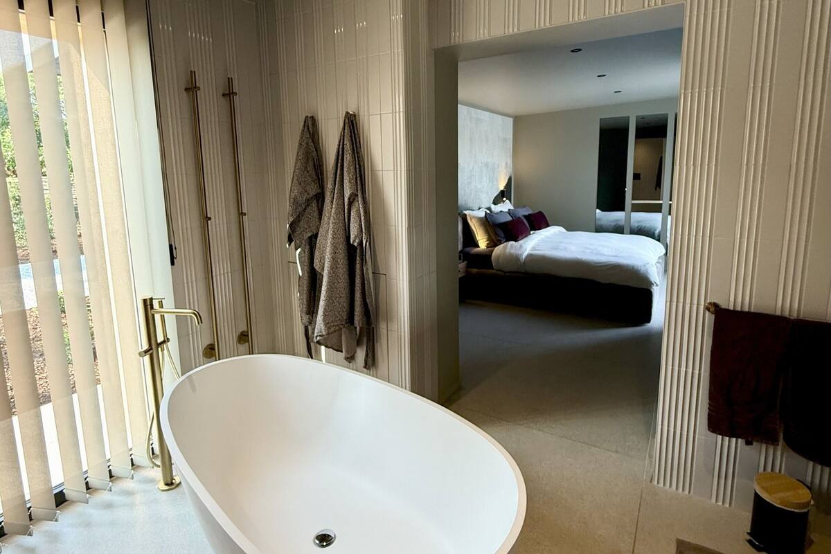 A modern bathroom with a freestanding bathtub, a shower area with vertical blinds, and a visible bedroom with a bed and nightstand.
