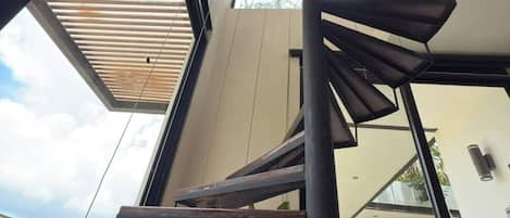 A spiral staircase with a wooden finish, leading to an upper floor with large windows and a view of greenery outside.