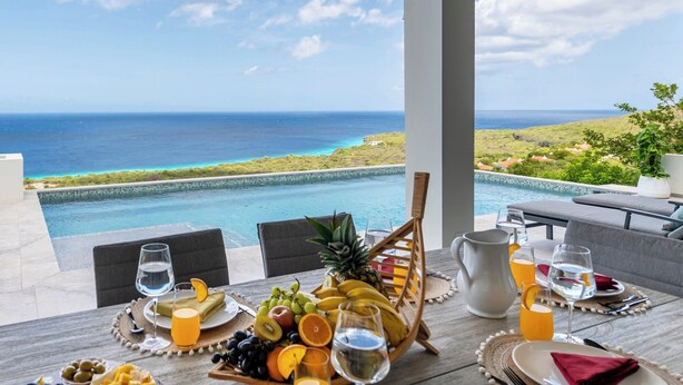 A dining table with fruit and drinks, overlooking a pool and ocean view.