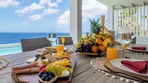 A dining table with a variety of fruits, cheeses, and beverages, overlooking a scenic ocean view.
