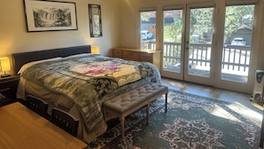 A bedroom with a bed, a bench, a desk, a nightstand, a ceiling fan, and a view through the glass doors.