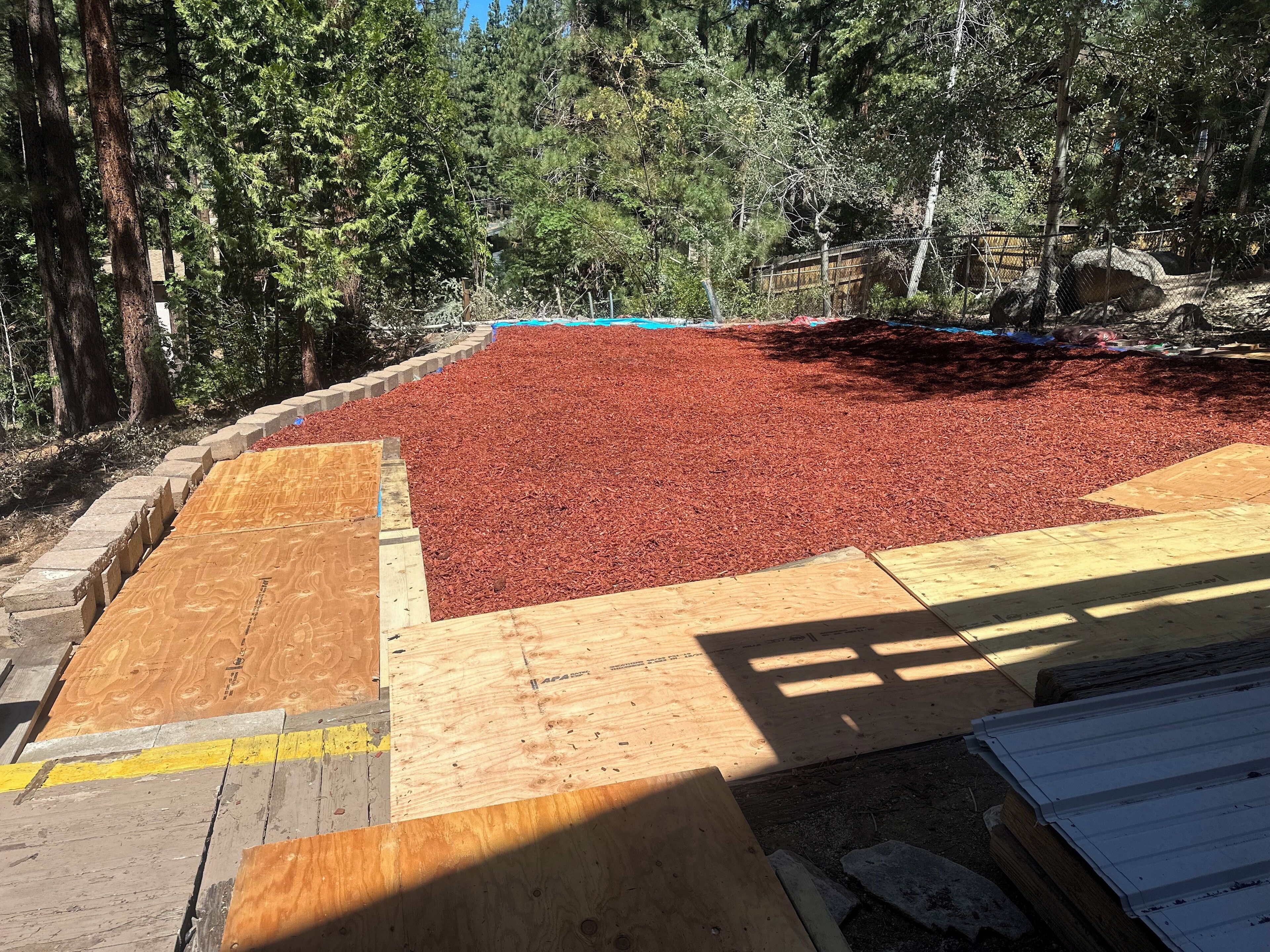 A construction site with a large area covered in red mulch, surrounded by trees and a wooden platform.