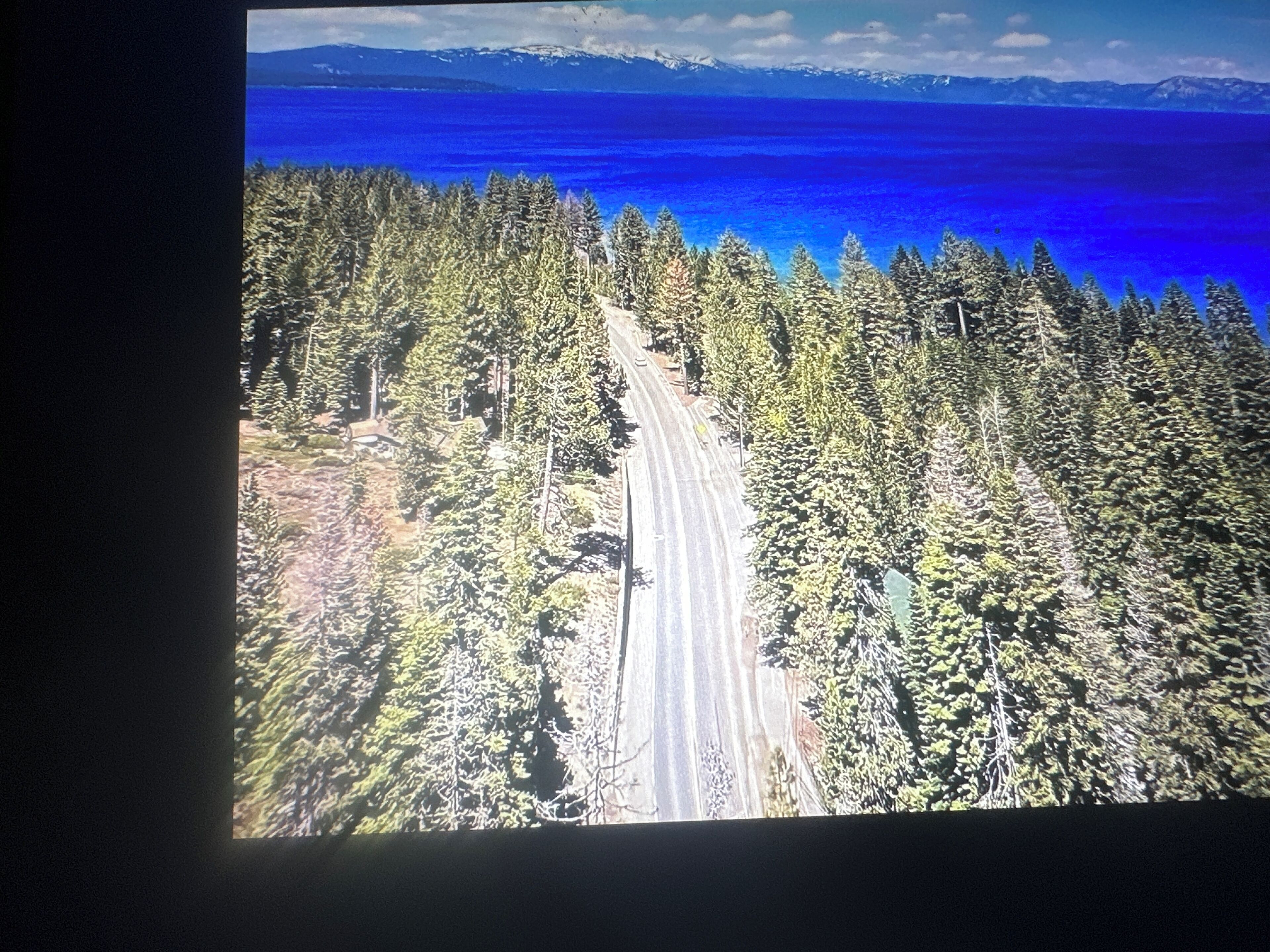 Vue aérienne d’une route traversant une forêt dense, avec un lac bleu clair et des montagnes en arrière-plan.