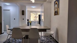 A dining area with a long table, white chairs, a vase with flowers, and a window with a view of buildings.