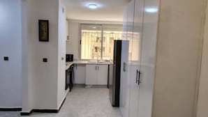 A narrow hallway leading to a kitchen with white cabinets, a black refrigerator, and a window with a view of buildings.