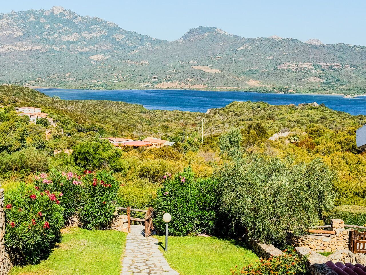 Paisagem costeira com um caminho de pedra que leva a um edifício com telhado vermelho, cercado por vegetação exuberante e vista para o mar e colinas.
