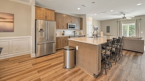 Una cocina moderna con armarios de madera, electrodomésticos de acero inoxidable, una isla central con taburetes y un amplio ventanal que proporciona luz natural.