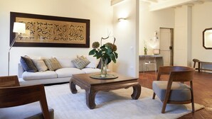 Ein Wohnzimmer mit einem weißen Sofa, einem Holztisch mit Blumenvase, einem Holzstuhl und einem großen abstrakten Gemälde an der Wand.