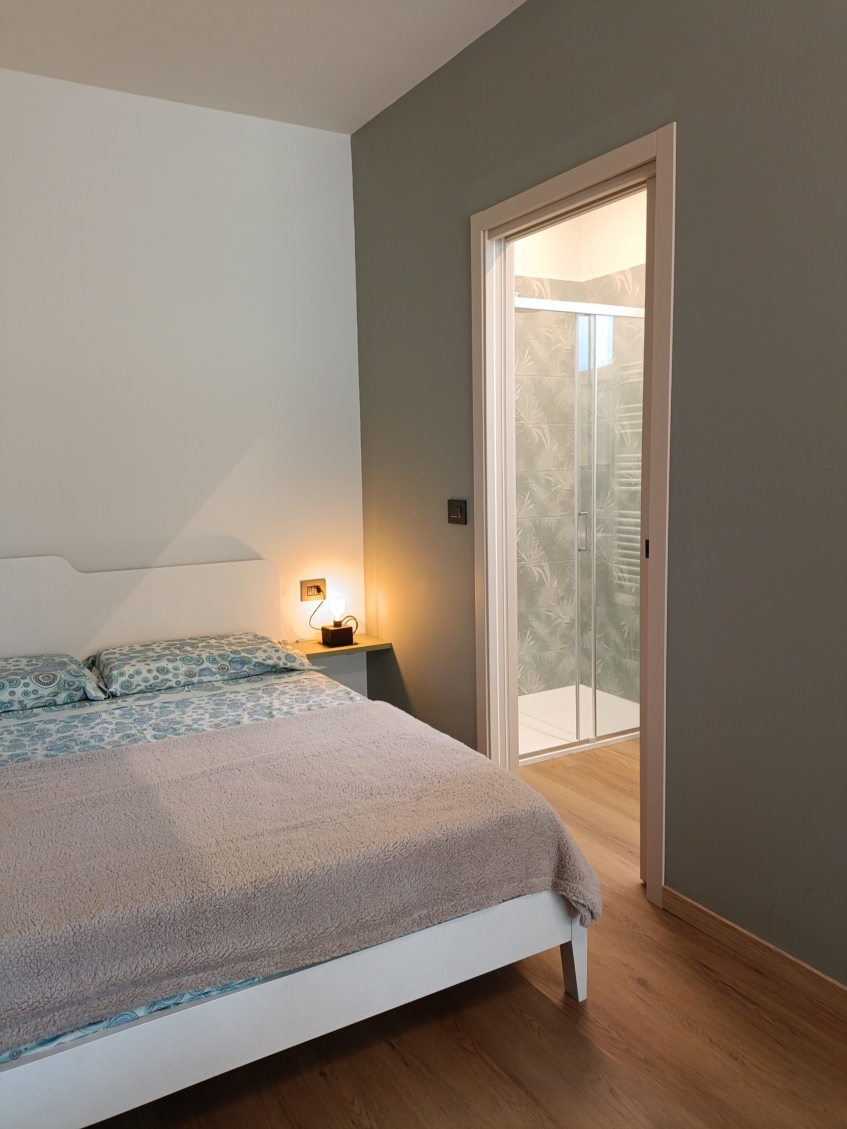 Een slaapkamer met een bed, nachtkastje, lamp en uitzicht op een balkon.