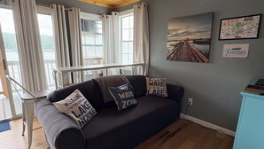 A cozy room with a dark blue sofa, wooden flooring, a ceiling with exposed beams, a window with curtains, and a wall-mounted air conditioner. - Charming 2-bedroom house boat in Norris Lake. (Speedwell)