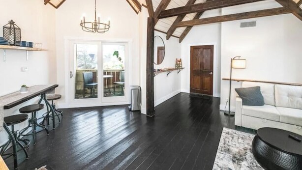 Una habitación amplia con techo alto, suelos de madera oscura, un sofá blanco, una zona de comedor con taburetes y un ventanal grande.