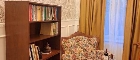 A room with a wooden bookshelf filled with books, a floral armchair, a table with a wine bottle and glass, and a patterned carpet.