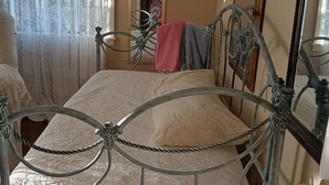 Une chambre avec un lit en métal, une couvre-lit en dentelle, un plancher en bois et un miroir au mur.
