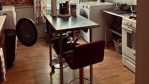 A kitchen with a brick wall, a window with floral curtains, a table with a chair, a washing machine, a stove, and a trash bin. - Charming 2-bedroom apartment in peaceful Warren with AC (Warren)