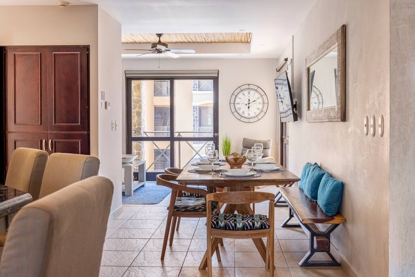 A dining area with a round table set for four, a clock on the wall, and a bench with cushions.