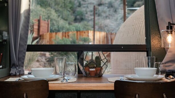 Un coin repas avec une table dressée pour deux personnes, une fenêtre avec des rideaux et une vue sur un pont et la verdure environnante.