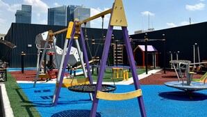 A playground with swings and climbing structures, surrounded by a fenced area with artificial turf and a view of tall buildings in the background. - Chaqar 198 (Tashkent)