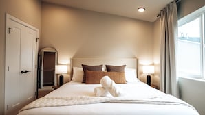 Una cama bien hecha con sábanas blancas y almohadas marrones, una lámpara de noche y un ventanal con cortinas.