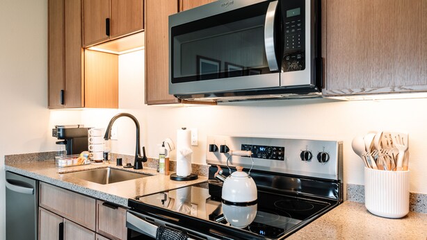 Una cocina moderna con microondas, horno y lavavajillas de acero inoxidable. Hay una encimera de granito, un hervidor y un portalapices.