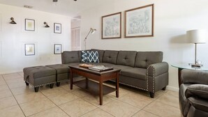 A living room with a sectional sofa, a coffee table, a ceiling fan, framed artwork on the walls, and a lamp on a side table. - Inviting home w/ big yard & walkable neighborhood! 3br/2ba (Tampa)