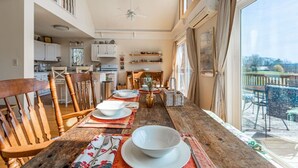 A dining area with wooden chairs and a table set for four, a kitchen in the background, and a large window with curtains.