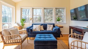 A cozy living room with a blue sofa, wicker chairs, a flat-screen TV, and large windows overlooking a wooded area.