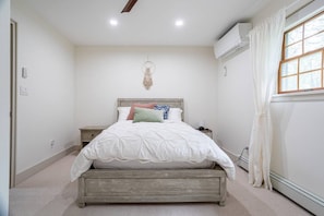 A bedroom with a bed, bedside table, wall-mounted air conditioning unit, and a window with curtains.