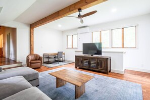 Un salon spacieux avec une poutre en bois au plafond, un téléviseur posé sur un meuble sombre en bois, un canapé en cuir, une table basse et une grande fenêtre laissant entrer la lumière naturelle.