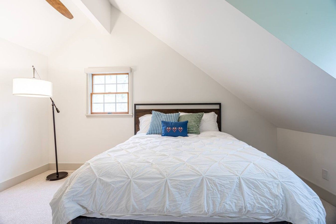 A bedroom with a sloped ceiling, a bed with a white quilt, a floor lamp, and a window.