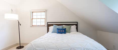 A bedroom with a sloped ceiling, a bed with a white quilt, a floor lamp, and a window.