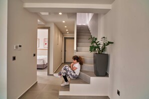 Una escalera con una planta en maceta, una persona sentada en los escalones, y un dormitorio visible al fondo.