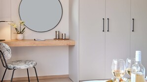 A modern room with a round mirror, a wooden shelf with decorative items, a chair, and a table with wine glasses and bottles.