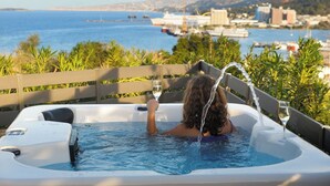 A person relaxing in a hot tub with a glass of water, overlooking a coastal cityscape.