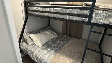 Ein Etagenbett mit gemustertem Bettbezug und Kissen, ein Fenster mit Vorhängen sowie ein kleiner Nachttisch mit Lampe.