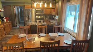 A dining area with a wooden table set for four, surrounded by wooden chairs. A kitchen with modern appliances and wooden cabinets is visible in the background. - 2 houses, huge yard, dog-friendly, close to ORV trails and lakes (St. Helen)