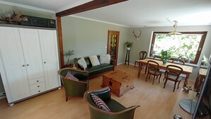 A spacious living room with a green sofa, wooden dining table, and chairs. There is a white cabinet and a wooden chest of drawers.